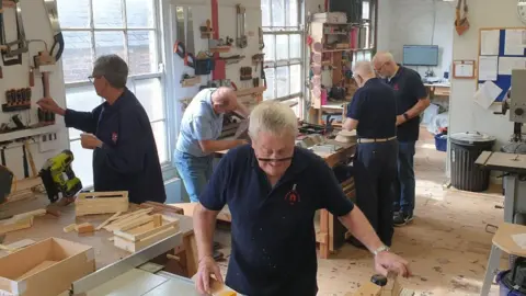 Ripon Men's Shed A woodworking workshop with five people actively engaged in various tasks. The space has large windows and overhead fluorescent lights. Tools like saws, clamps, and measuring instruments are neatly arranged on wall racks.