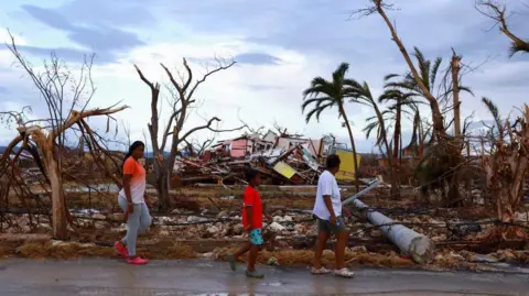 Three people walk past damaged houses and fallen trees. The rubble is piled up behind them as they walk from left to right. The sky is light blue with clouds. 