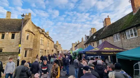 Lots of people walking around Oundle's market in a narrow street, lined with sandstone two-storey houses, with people looking at the stalls. There are various canopies of different colours