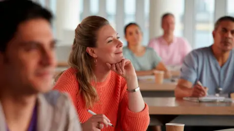 A stock image of a group of mature learners in a classroom. A blonde long-haired woman in an orange jumper is in focus, her chin resting on one hand and a pen in the other. She is smiling as she looks to the front. Three men and a woman sit on surrounding tables, out of focus.