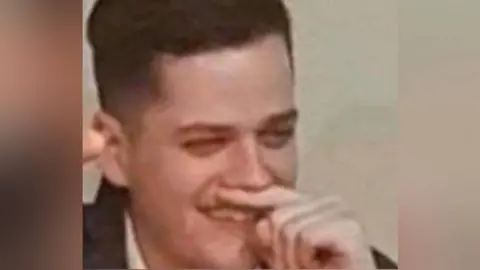 A man holds his finger under his nose while holding his right arm in the air