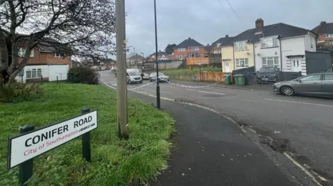 A shot of Conifer Road. Police tape can be seen blocking off access to the street.