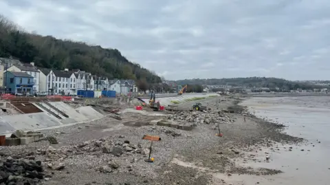 Mumbles beach, with lots of rubble and machines working near the shore. Buildings are above the shoreline next to the road 