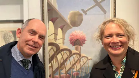 Paul and Helen smile at the camera - they are both wearing smart coats. They are in front of one of Orlik's paintings. It is surrealist but very accurate - they're is a stylised plane flying over a ocean and a bridge, with a tower and large flower on top.