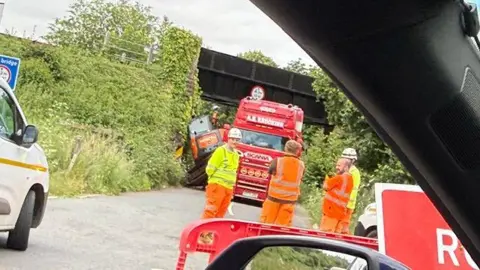 A red lorry stuck beneath a railway bridge. An orange digger has fallen from one side of the vehicle and is leaning against an embankment. a group of men in high-vis jackets stand in front of the vehicles. 