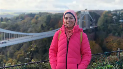 Dr Norah El-Gohary Dr Norah El-Gohary in a pink coat standing in front of Clifton Suspension Bridge.