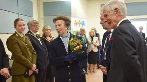 Princess Anne from NHS Somerset speaks to people standing to welcome her in a large hall. She is wearing flowers.