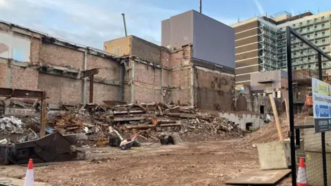 Alex Pope/BBC A pile of rubble from a demolished building, with a block of flats in the background. A metal fence is to the left and two traffic cones to the left and right.