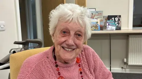 An older woman, with short white hair smiles at the camera. She wears a pink knitted jumper and a long necklace with different shaped pink and purple beads.