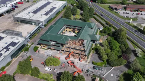 Eddie Mitchell An aerial image of demolition works on a square building with a dark green roof. The building is surrounded by cranes and has a chunk taken out of its side. the roof has a number of triangular layers.
