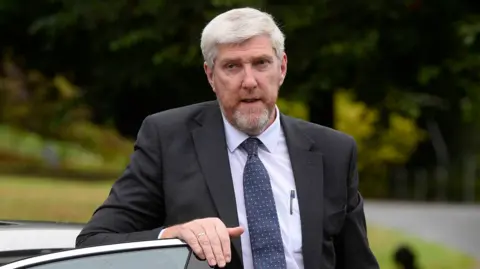 PA Media John O'Dowd - A grey-haired man wearing a dark suit jacket, a light-coloured collared shirt and a pattern tie. He is resting his hand on top of a car door with blurred shrubbery in the background.