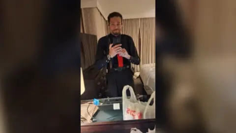 Cole Tomas Allen in a selfie taken in a mirror. He's wearing a black shirt and trousers and a red tie.