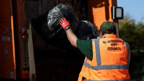 Reuters A man wearing an orange hi-vis vest which says "Birmingham City Council" on the back, throws two black bags of rubbish into the back of a bin lorry.