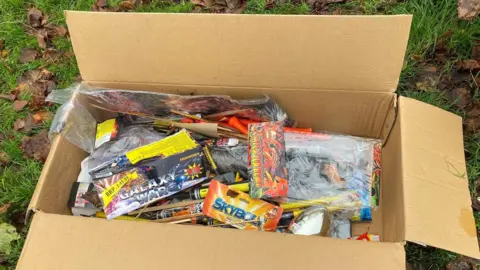 A cardboard filled with brightly packaged fireworks, all with names like Galaxy War. The box is on grass with leaves around it.