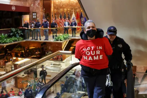 A woman in a red top that reads 'not in our name' has her hand behind her back and is being escorted up an escalator by a police officer 