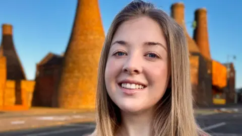 Stoke-on-Trent city council Heather Blurton stands in front of bottle kilns, she has long blonde, straight hair and is smiling at the camera. 