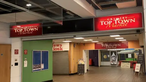 Lara King / BBC The entrance to Top Town Market. There are three signs overhead bearing the market's name.  A News Kiosk sign can also be seen. The kiosk has its shutters down. 