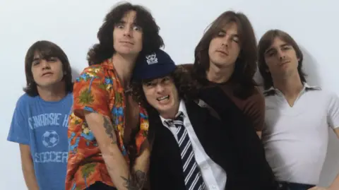Getty Images AC/DC posing in a photoshoot picture. Angus Young is in the entre wearing a school uniform and cap. 