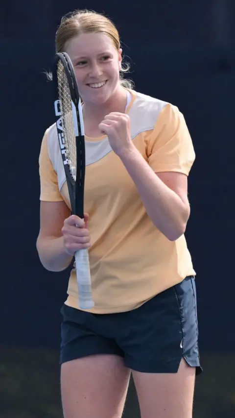 Daniella Britton stands on a tennis court with one hand lifted in a triumphant fist. She holds a tennis racket in her other hand. Daniella wears a pale orange short sleeved top with white detailing and navy blue shorts. She has ginger hair tied back in a low ponytail.
