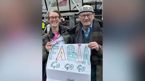 Jim Pennington Fabia and her dad, smiling to camera holding up a banner, saying 'Fabia, go, go, go'. She is holding up her medal at the end of the race