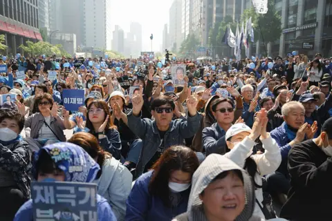 Getty Images A large crowd of Lee Jae-myung supporters gathers in a city street lined with tall, modern buildings, some of them holding blue placards