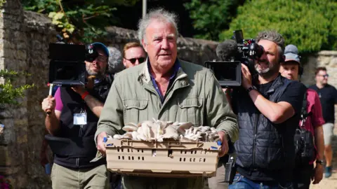 Jeremy Clarkson, reporter mediów PA, ma na sobie zielony płaszcz i niesie tacę z grzybami. Za nim idzie ekipa filmowa