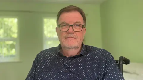 Mid-shot of Ron Patton, dark hair, glasses and a beard. He is wearing a black polka-dot shirt.