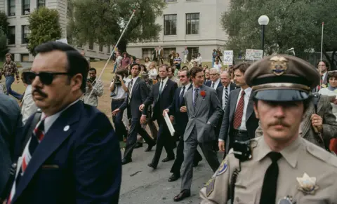 Michael Ochs Archives / Getty Images Prince Charles walks through the campus with security and protesters carrying signs with camera operating journalists in the background.