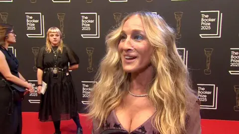 Sarah Jessica Parker, with long wavy blonde hair and a pale purple dress, smiles on the red carpet.