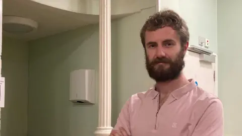Emma Howgego/BBC A man stares at the camera with his arms folded. He has a beard and wears a light pink top. In the background is a green wall with a white pillar and fire door. A food menu is on the wall to the right and some cereal boxes are to the left. 