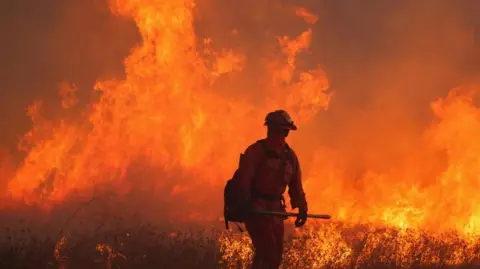 Reuters Firefighter mengenakan pakaian pelindung penuh, termasuk topeng dan helm, dengan api besar menutupi area di belakangnya.