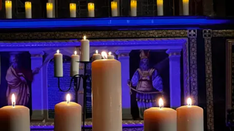 Neil Fisher A close up of several large candles burning in front of an altar-type scene, with subtle blue lighting 