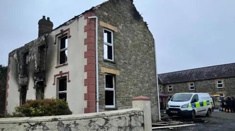 House badly damaged by fire, with scorch marks on the exterior walls and from windows and the door. A police van is parked behind, with emergency services officers nearby