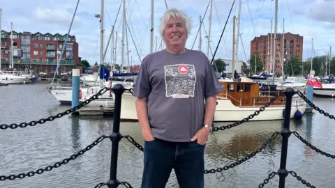 A man with a mop of grey hair smiles as he stands in front of a marina with boats behind him. He is wearing a grey T-shirt with waves design and blue jeans.