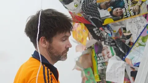 Kaku San Stuart Langley is working on a piece of art which involves crumpled up magazines and other bits of paper placed on a wall. He has short brown hair and a beard, and is wearing a orange and blue zipped sweater.