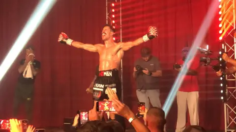 A boxer dressed in black shorts with sponsor branding, his arms outstretched with red, black and white boxing gloves on his hands. He is standing on a stage in front of a red curtain, surrounded by photographers, fans taking pictures on their phones and stage lights on steel-lattice towers. 