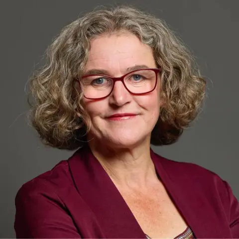 Jodie Gosling A woman stands against a grey background. She has curly grey hair and glasses. She is wearing a maroon jacket.