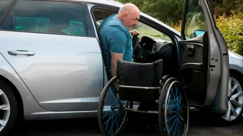 Getty Images A man is in the middle of getting from a wheelchair into the driver's seat of a car - we can see the driver's side door open, where the wheelchair is sitting empty. The man is mostly in the seat with some of his head still leaning out the door. The car is a silver estate, and behind where it is parked is a green bush.
