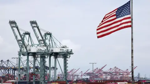 An American flag flies in front of shipping containers and cranes at a port.