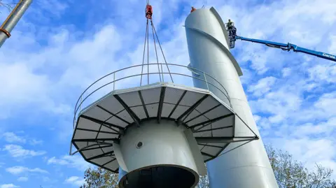 Cranfield University The construction of a new radar tower with a crane lifting a section on to the top of the tower. There is blue sky and the tops of trees are in the background.