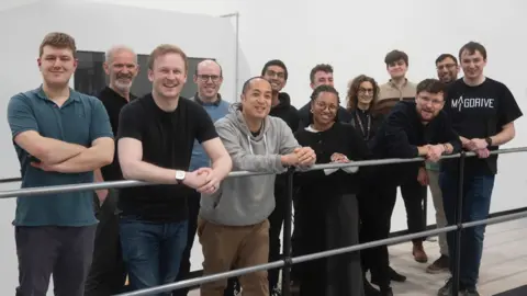 Eleven men and two women standing by a rail, dressed in black or navy against a white wall. they are smiling at the camera.