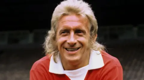 PA Media Denis Law, with a blonde mullet, in his early 1970s Manchester United red shirt with extended white V-necked collar