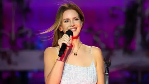 Getty Images Lana Del Rey in a white dress holding a microphone