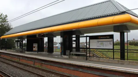 Meadow Well station which is a train platform which is partially covered by a grey and yellow roof. There is a board on the platform with a map and an electronic board with departures on it. The back of the station is open and a grassy bank can be seen with houses in the background.