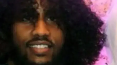 A zoomed in image of a man's head, smiling at the camera. He has a black beard and moustache and long curly black hair. 