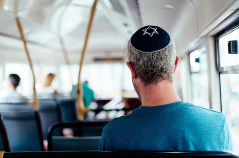 Colour image depicting a Jewish man wearing a traditional Jewish skull cap with star of David design on a bus. Other passengers are defocused in the background.