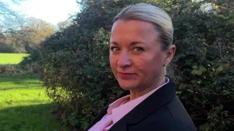 Lucy Edgeworth, wearing a pink striped shirt with collars and a dark jacket over the top, stands looking sideways at the camera. She has blonde hair tied up in a bun. Behind her are bushes and a grassy area