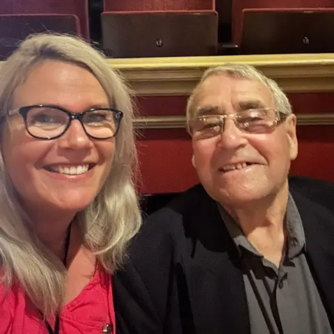 Rachel Firth Woman with long blonde hair and glasses and man with short hair and glasses. They are both smiling. She is wearing a red top and he is wearing a black jacket and a grey shirt. They are in front of a row of red theatre style fabric folding seats.