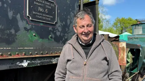 A man wearing a grey fleece top is standing alongside a black railway engine which has flaky paintwork. Behind him is a green railway engine which appears to be in a state of disrepair. There are trees behind that.