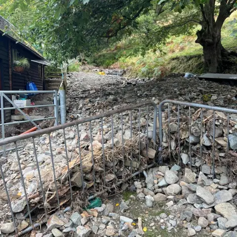 A metal fence with stones and boulders piled up on the other side, in the background there is a path/road, and a grassy slope with a tree on it